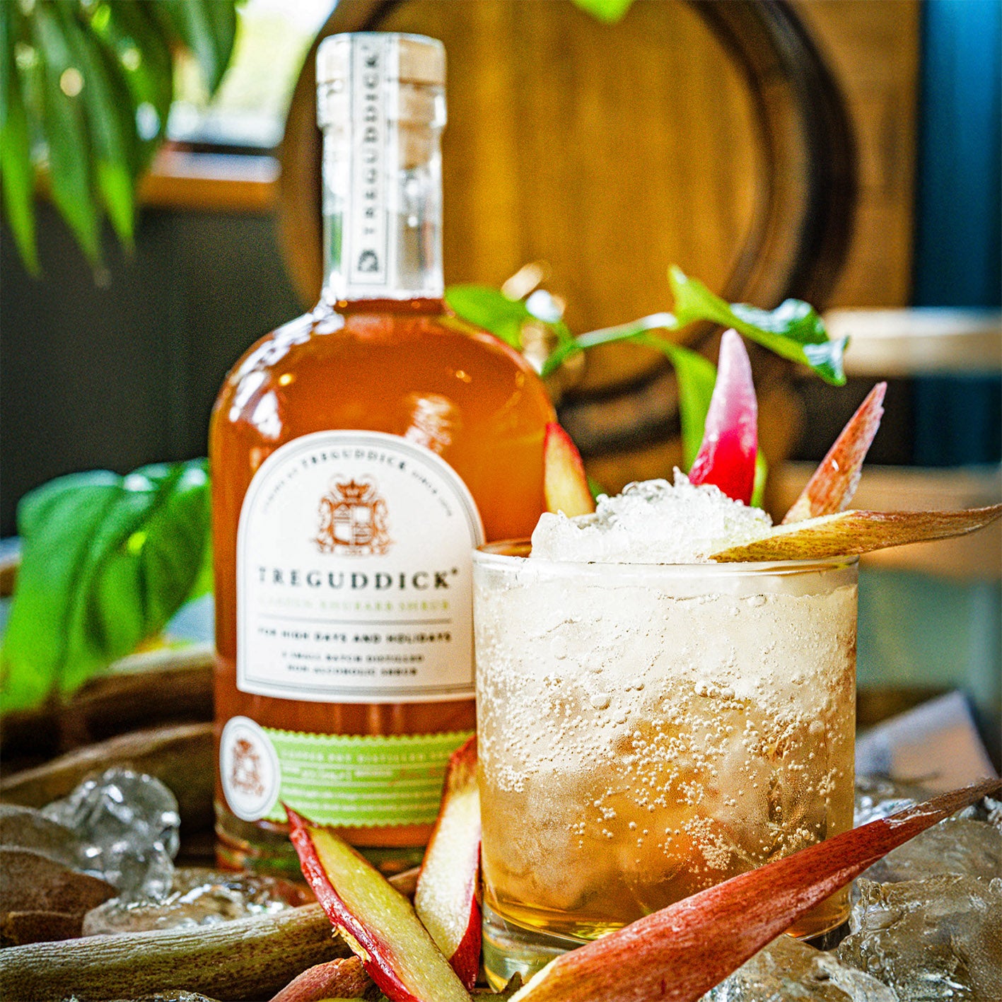 Bottle of Treguddick gin with a cocktail garnished with apples and ice, set against a wooden barrel background.